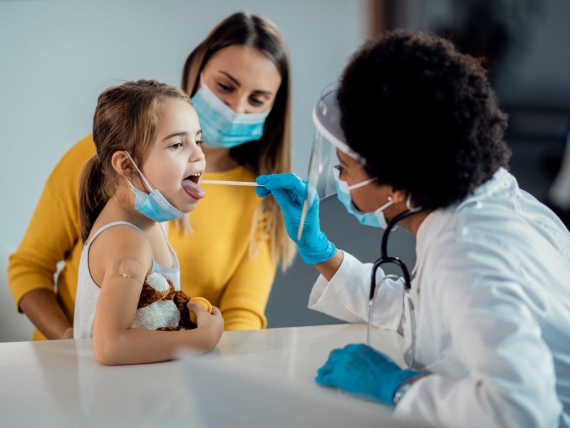 African American pediatrician examining throat of a small girl who came with mother to medical appointment during coronavirus pandemic. Focus is on girl.